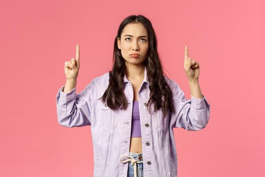 Portrait of gloomy and displeased, young frowning girl in denim jacket, grimacing looking and pointing fingers up disappointed, express regret or jealousy, standing pink background.
