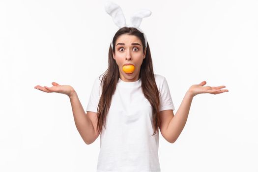 Portrait of confused and indeisive young funny woman with rabbit ears, holding colored Easter egg in mouth, staring clueless camera with popped eyes, shrugging and spread hands sideways.