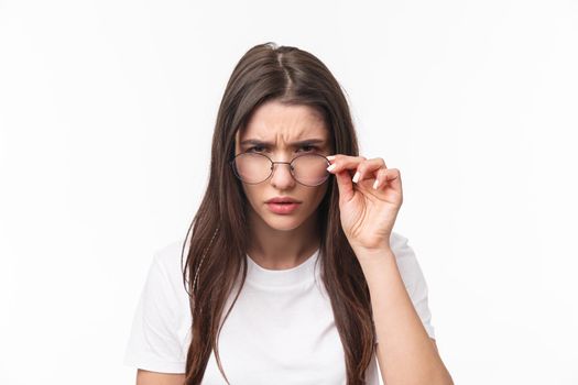 Close-up portrait of suspicious young serious-looking woman, look from under glasses, squinting at person with judgemental disbelief stare, standing white background, have doubts.