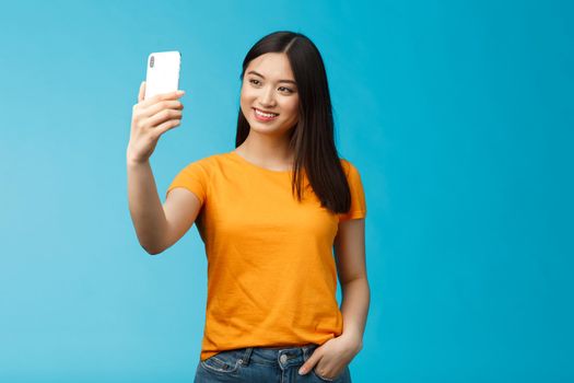 Stylish good-looking asian cute girl hold smartphone talking video-call smiling broadly look phone screen, taking selfie, feeling pretty, talking followers online social media, blue background.