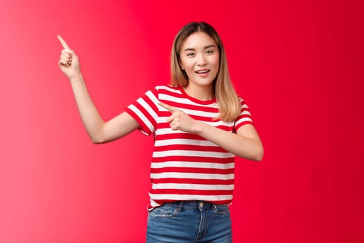 Enthusiastic friendly modern asian blond girl suggest perfect place hang promo pointing upper left corner smiling look camera assertive introduce project, recommend link, stand red background.