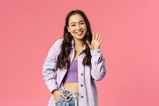 Hello nice to meet you. Cute stylish young girl meet new people at work, waving hand and smiling with little laugh as greeting, saying hi, welcoming newbies, standing pink background.