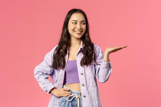 Girl presents you something worth your attention. Portrait of cheerful attractive asian female introduce product, pointing with palm and looking right, smiling pleased, pink background.