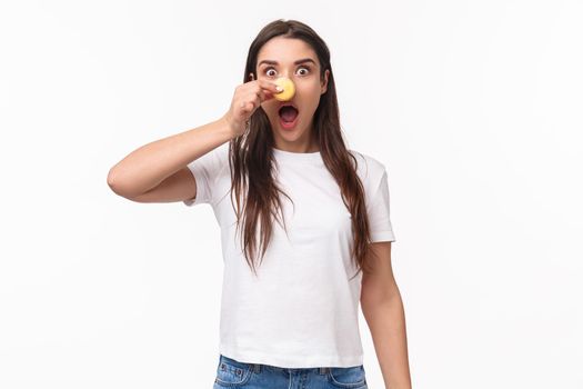 Portrait of funny adorable young brunette woman in t-shirt, open mouth excited, look enthusiastic, joking making nose from macaron and staring camera, eating delicious desserts, white background.