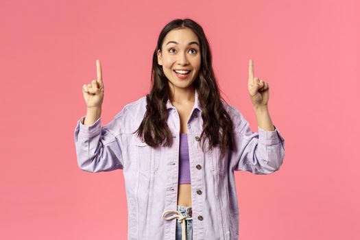 Enthusiastic young pretty girl looking and pointing fingers up with happy smile, see promo, interested in taking part event, party, found great online courses, standing pink background.