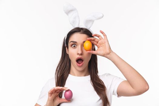 Easter, holidays and spring concept. Close-up portrait of amused, enthusiastic young woman in rabbit ears, stare camera with opened mouth, holding colored egg over eye and in hand.