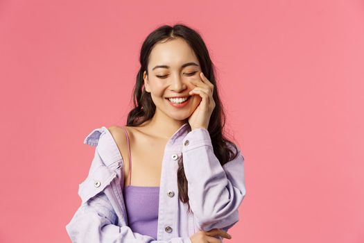 Lifestyle, fashion and beauty concept. Close-up portrait of cheerful, lovely young asian woman close eyes, enjoying day, laughing happily, touch clean skin, pink background.