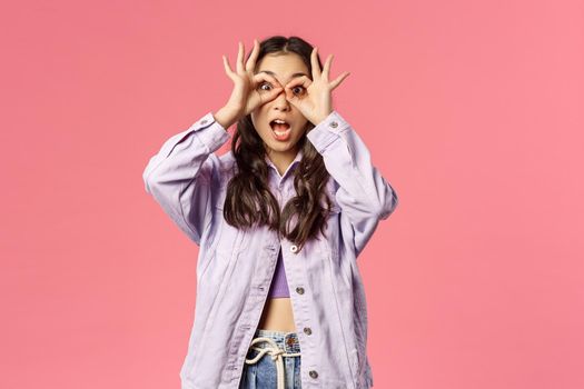 Portrait of funny and silly, playful stylish girl making funny expressions and grimaces, hold hands over eyes like glases or mask, fool around like child, standing pink background.