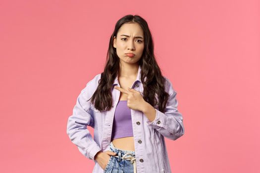 Portrait of moody, upset young asian girl, pouting and frowning disappointed, pointing finger left, jealous of not having something she wants, feel offended or uneasy, pink background.