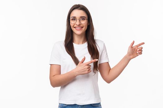 Waist-up portrait of enthusiastic, pleasant brunette woman in glasses inviting check out promo, visit or click link, pointing fingers right and smiling at camera, standing white background.
