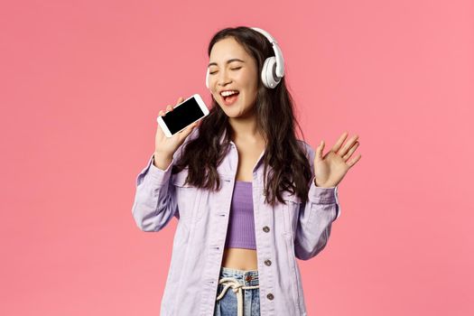 Waist-up portrait of stylish happy woman using headphones to listen awesome new track, download karaoke app and using smartphone like microphone, singing along awesome music.