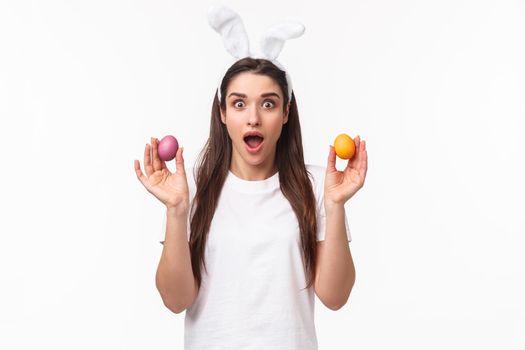 Portrait of surprised and amazed young brunette girl holding two colored eggs, wearing rabbit ears, party outfit as celebrating Easter day, enjoying holy holiday, white background.