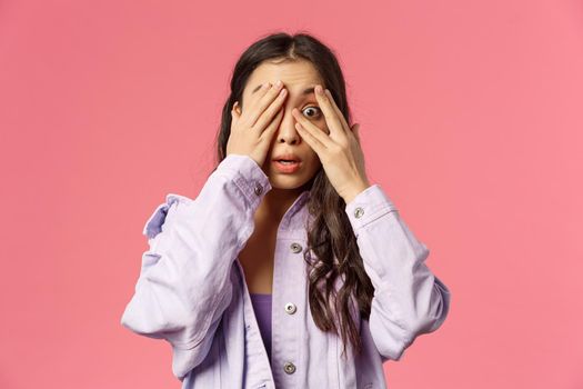 Close-up portrait of scared and alarmed young asian woman panicking, cover eyes from fear, peeking with one eye at something shocking and embarrassing, stand pink background.