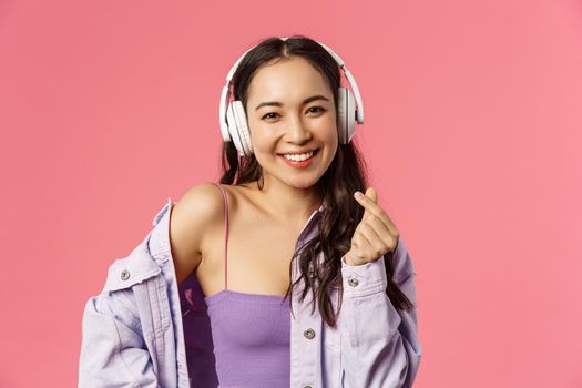 Close-up portrait of attractive happy young modern asian girl, listening music in headphones, show korean heart sign, enjoying favorite music or podcast, bought earphones with discount.