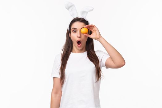 Waist-up portrait of amazed and surprised young woman seeing something awesome, hold colored egg over eye, staring shocked camera, gasping impressed, wear rabbit ears, celebrate Easter day.
