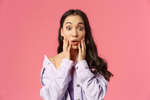 Close-up portrait of surprised and amazed, gasping young asian woman cant believe something astounding happened, hold hands on cheeks, gasping and staring astonished, pink background.