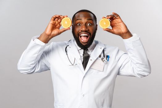 Healthcare, healthy diet and disease concept. Excited handsome african-american doctor in white coat, showing pieces of oranges and smiling amused, advice eat more food consist vitamin c, fruits.