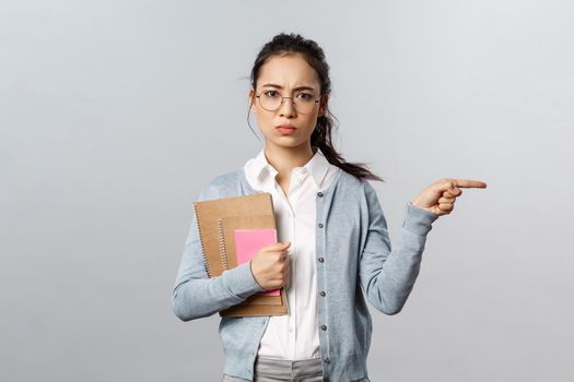 Education, teachers, university and schools concept. Go there, leave now. Serious-looking disappointed frowning female tutor look displeased, pointing finger right, standing grey background.