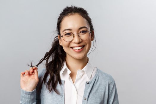 Close-up portrait of flirty, enthusiastic brunette asian female in glasses, beaming smile, roll strand of hair between fingers as coquettish talking to person she likes, standing grey background.