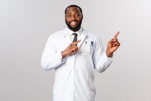 Friendly handsome african-american male doctor explaining main symptoms covid19, showing chart or prescription how be safe, pointing upper right corner and smiling, recommend use face mask.