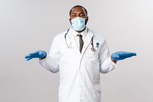 Covid-19, coronavirus patient treatment and disease concept. Tired and bothered african-american doctor in face mask and latex gloves, standing with hands spread sideways prepare for usual check-up.