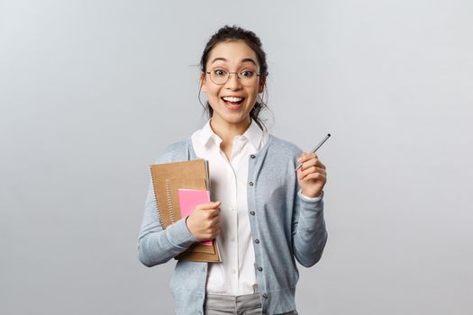 Education, teachers, university and schools concept. Enthusiastic young asian female in glasses, holding pencil and notebooks, write good mark at student essay, pleased with great homework.