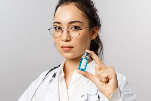 Close-up portrait of serious-looking young asian infectionist, female doctor showing ampule with coronavirus vaccine, invented covid19 medicine, look determined, start treating patients.