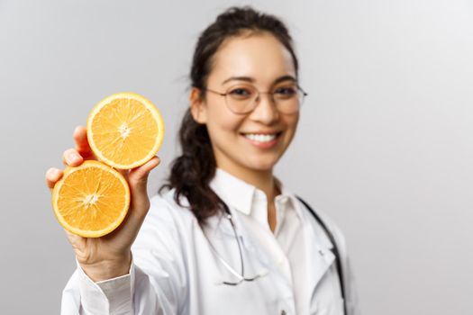 Close-up portrait of asian female doctor, therapist showing pieces of orange and smiling, give advice eating healthy food, more vitamins from fruits, standing grey background.