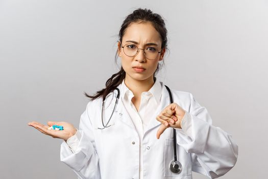 Healthcare, medicine and pharmacy concept. Portrait of worried asian female doctor, frowning disturbed, thumbs-down and hold bad pills, advice not to self-medicate from virus, contact your therapist.