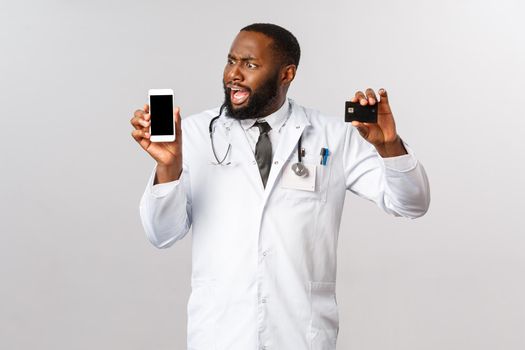 Covid19, pandemic and online medicine concept. Complaining angry african-american man looking at mobile phone display, hold credit card, outraged with huge bills and expensive app subscribtion.