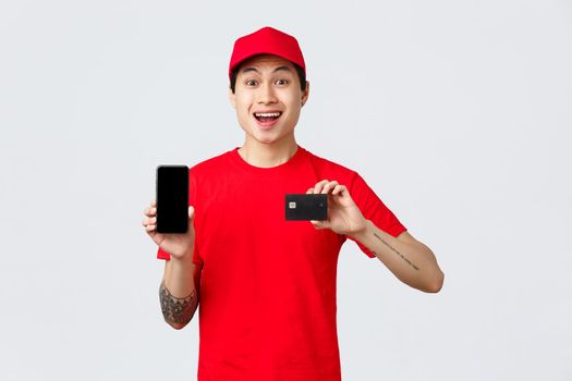 Delivery application, online orders and shipping concept. Quarantine wont stop from shopping. Cheerful asian courier in red cap and t-shirt, showing smartphone display and credit card.