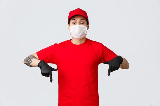 Amazed and excited asian delivery guy in red uniform cap and t-shirt express his shock and emotions with eyes, pointing fingers down in protective gloves and medical mask, showing new promo offer.