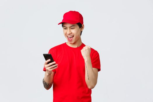 Happy young asian delivery guy in red uniform t-shirt and cap, reading good news smartphone screen, make fist pump chanting great bonuses or success. Courier say yes, tracking orders.