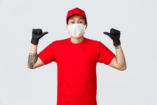 Cheerful, happy asian delivery guy in red cap and t-shirt, wearing protective gloves and medical mask, smiling eyes and pointing himself, got work in delivery company, become employee of year.