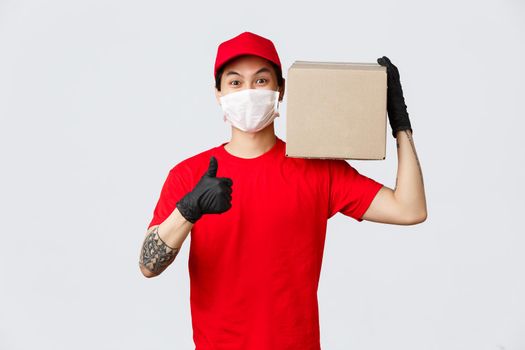 Concept of delivery and carriers during coronavirus pandemic. Smiling asian courier in red cap, t-shirt, wearing medical mask and gloves, covid-19 safety measures on work, hold package, make thumb-up.
