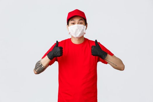 Sounds good. Cheerful asian delivery man in red cap and t-shirt, company uniform and medical gloves, protective mask, thumbs-up, say yes, approve or agree, recommend courier service.