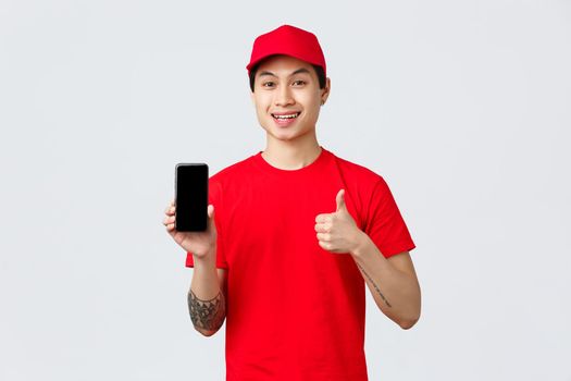 Delivery application, online shopping and shipping concept. Handsome asian courier in red t-shirt and cap, show thumb-up in approval and mobile phone screen app, provide fast and quality service.
