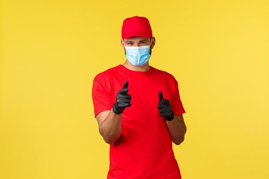Delivery during pandemic, covid-19, safe shipping, online shopping concept. Friendly pleasant courier in red uniform, medical mask and gloves pointing cheeky camera, promote express mail.