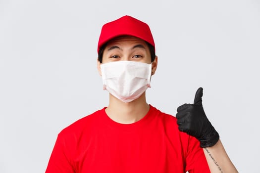 Covid-19, self-quarantine online shopping and delivery concept. Cheerful asian courier in medical mask and protective gloves, wear red uniform, show thumb-up in approval, recommend make safe orders.