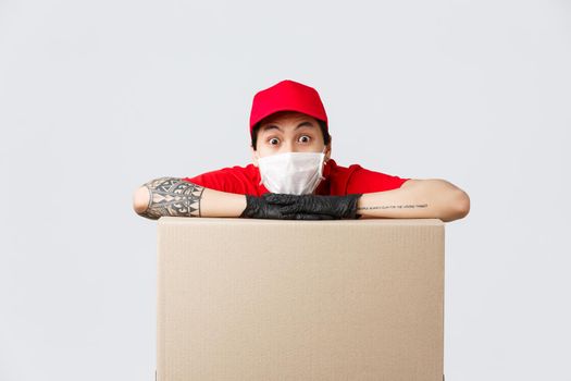 Excited asian delivery guy in medical mask and protective gloves, leaning on box parcel and staring camera astonished. Courier working during coronavirus pandemic, safely deliver orders.