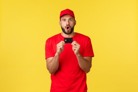 Contactless payment, online delivery, internet shopping concept. Excited and amazed young male employee in company uniform, red t-shirt and cap telling amazing news, show credit card.