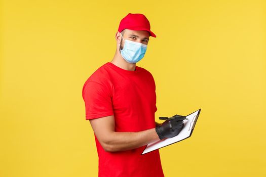Express delivery during pandemic, covid-19, safe shipping, online shopping concept. Pleasant young courier in red uniform and medical mask, fill in order on clipboard, look camera.