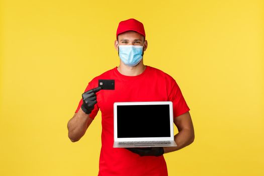 covid-19, safe shopping, express delivery and order tracking concept. Smiling courier in medical mask, uniform, showing credit card and webpage on laptop screen, contactless easy payment.