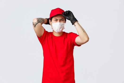 Reluctant asian delivery guy put on red cap, frowning and grimacing displeased, wear protective gloves and medical mask, unwilling carry heavy box. Courier complaining hard work.