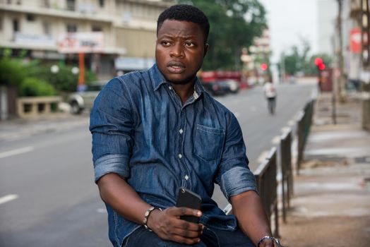 Confident businessman or manager sitting on an iron bar, holding a cell phone and looking aside in the city
