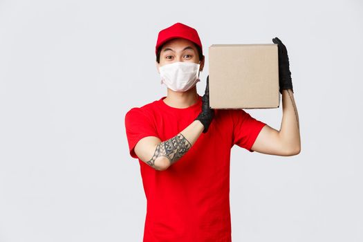 Concept of delivery during coronavirus pandemic. Charismatic asian courier holding package on shoulder, wear protective gloves and medical mask for client safety, give client their order.