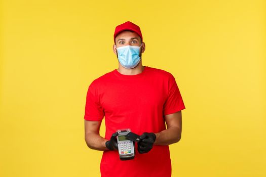 Food delivery, tracking orders, covid-19 and self-quarantine concept. Excited and amused courier in red uniform, medical mask and gloves, stare camera, using POS terminal and credit card.
