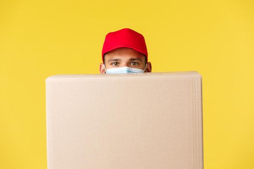 Express delivery during pandemic, covid-19, safe shipping, online shopping concept. Funny courier in red uniform cap, hiding behind package box, wear medical mask, yellow background.