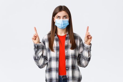 Coronavirus outbreak, leisure on quarantine, social distancing and emotions concept. Interested serious young woman in medical mask and casual outfit, looking and pointing up banner.