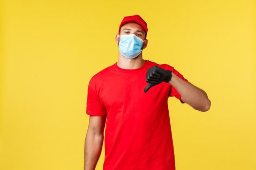 Express delivery during pandemic, covid-19, safe shipping, online shopping concept. Unamused young reluctant courier in medical mask and uniform, show thumb-down disappointed or displeased.